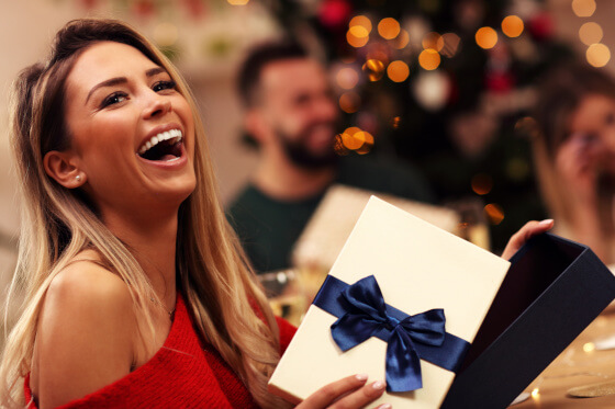 Zu sehen ist eine junge Frau, die ein Geschenk auspackt und sich sehr freut