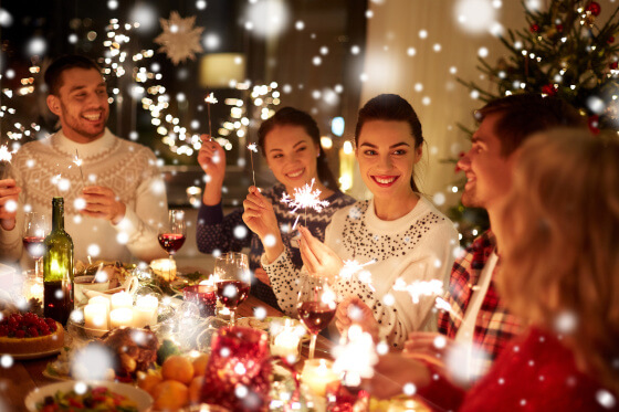 Zu sehen ist eine Gruppe von Leuten, die gemeinsam am Tisch sitzen und  eine Weihnachtsfeier feiern. Es ist festlich geschmückt und in den Händen halten einige eine Wunderkerze